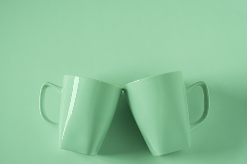 2 monochromatic green coffee mugs on green background clinking in cheers with blank empty room space for text, copy, or copyspace. Modern top view concept of two cups with solid background backdrop.