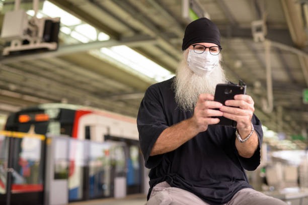 istock Mature bearded hipster man using phone with mask for protection from corona virus outbreak at train station 1222138348
