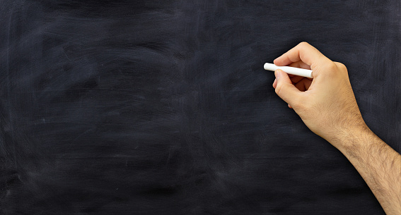 Man hand with a chalk isolated on black background. Teacher holding a white chalk against a chalkboard, copy space