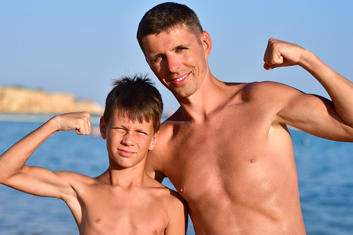 Son and father show muscles at sea.