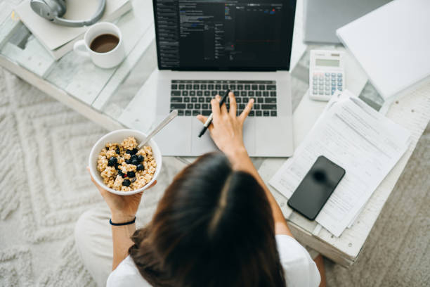 Overhead view of busy young Asian woman having breakfast while working from home using laptop computer and handling paperworks in the living room at home