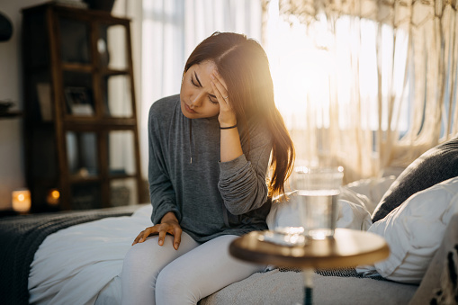 Young Asian woman sitting on the bed feeling sick and suffering from headache, a glass of water and medicine on the side table