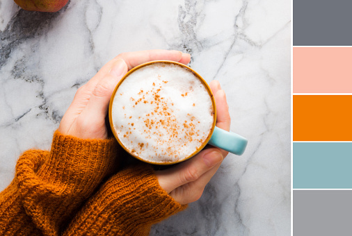 Autumn moody background with mug of latte coffee and pumpkins on marble table. Flat lay in fall colors. Female hands in cozy sweater. Color swatch