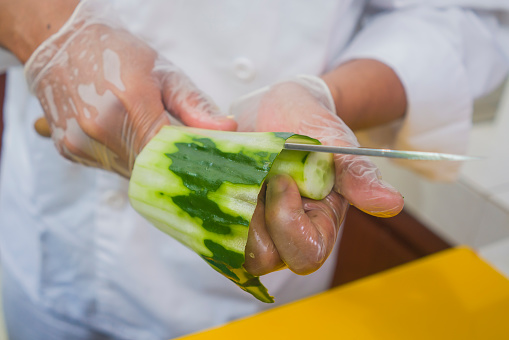hands in gloves of Asian professional chef preparing Japanese sushi rolls cutting cucumber with knife at home or restaurant kitchen making traditional Japan food
