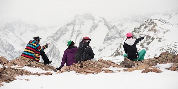 Group of female and male snowboarder and skier, sit and rest on the rock in the snow, and enjoy in beautiful landscape of panoramic Alps mountain range viewpoint. Back view. French Alps, Les 2 Alpes