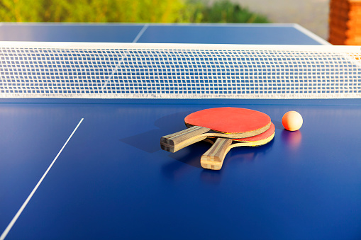 From above of similar red ping pong paddles and small ball on blue table on sunny summer day