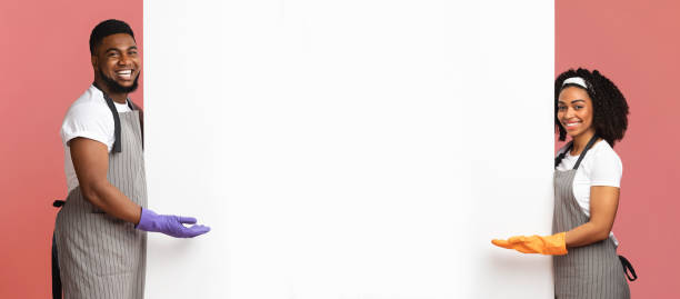 Spring Cleaning Ad. Positive African Couple In Rubber Gloves And Aprons Pointing At White Advertisement Board With Copy Space For Your Text, Panorama