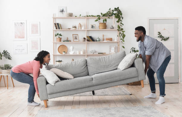 Black couple moving sofa in living room, replacing furniture at home Happy african american couple moving sofa in living room, replacing furniture at home, side view arranging stock pictures, royalty-free photos & images