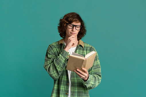 Back to school concept. Industrious student in glasses reading interesting book on turquoise background
