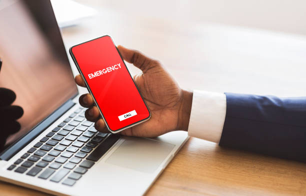 Worker holding smart phone with emergency number Safety Concept. Male hand holding smart phone with emergency number on red screen over office background and laptop disaster stock pictures, royalty-free photos & images