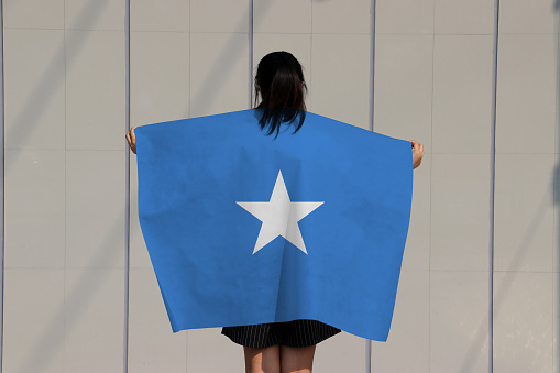 Woman is holding Somalia flag on her shoulder and turn back on grey background.