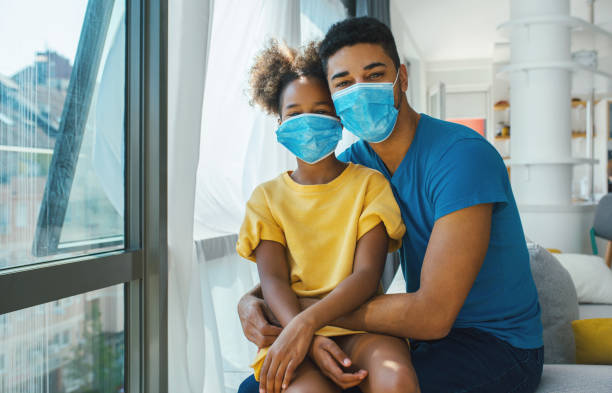 Father and daughter sitting by a window during coronavirus quarantine. Closeup front view of a mid 30's african american man with his daughter staying at home during coronavirus pandemic. They are staying at home, wearing face masks and looking at the camera. corona sun photos stock pictures, royalty-free photos & images