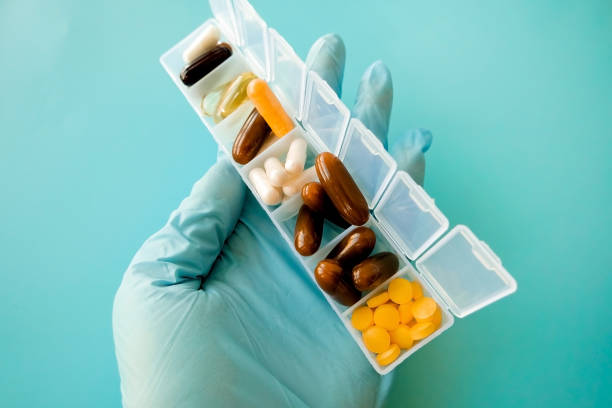 uma mão com uma luva médica contém cápsulas multicoloridas, uma caixa de comprimidos com medicamentos em um fundo azul - vitamin pill lecithin group of objects capsule - fotografias e filmes do acervo