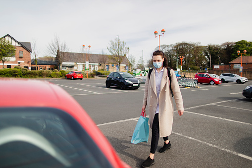 A female healthcare worker outside walking on a parking lot after buying groceries. She is holding a carrier bag and walking to her car while wearing a protective face mask.