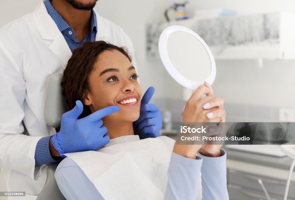 Attractive lady checking her beautiful smile in mirror Attractive black lady checking her beautiful smile in mirror after stomatological treatment Dentist Stock Photo