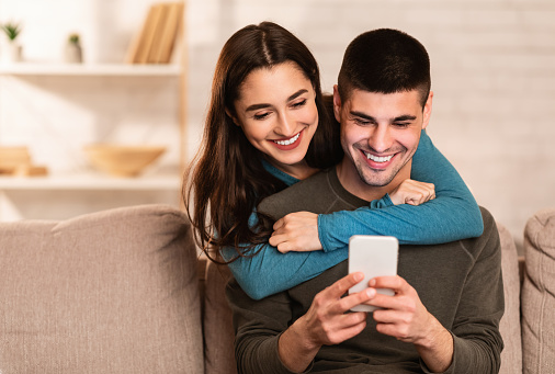 Romantic millennial couple sharing mobile sitting on couch at their apartment, girl hugging her man from the back