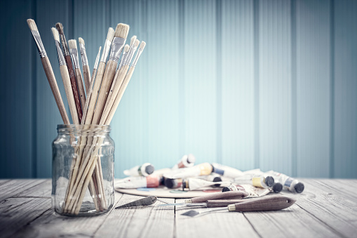 Paint brushes in a glass jar with paints and palette art and craft background