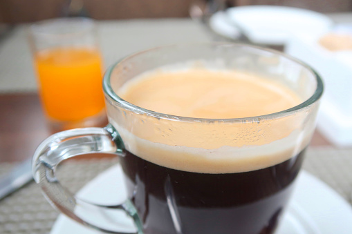 Photo showing a freshly made, hot cup of Americano (black) coffee, pictured on a restaurant table The coffee is presented in a simple, stylish glass mug with a blurred back ground of breakfast paraphernalia.