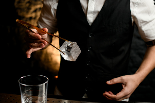 Close-up man at bar accurate holding large cube of ice with tweezers in his hand.