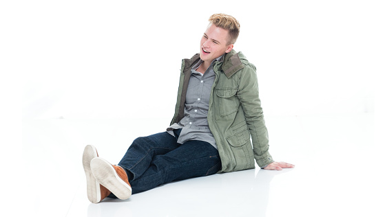 Profile view of aged 18-19 years old caucasian young male sitting on floor in front of white background wearing button down shirt who is happy