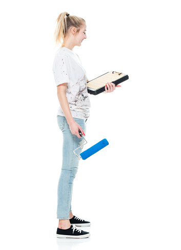 Side view of aged 18-19 years old who is beautiful with blond hair caucasian female house painter standing in front of white background wearing canvas shoe who is cheerful who is painting and holding checklist