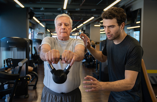 Senior man exercising at the gym with assistance from a personal trainer and using a kettlebell