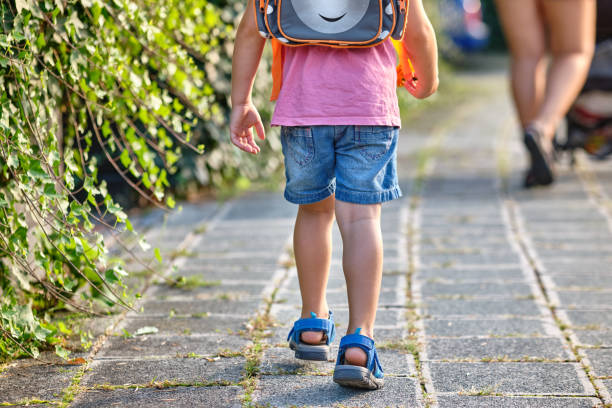 Rear view of 3 year old child walking behind its mother with rucksack Rear view of a 3 year old child walking behind his mother with a rucksack on the sidewalk on a sunny summer day. Seen in Germany in August. young hot stock pictures, royalty-free photos & images