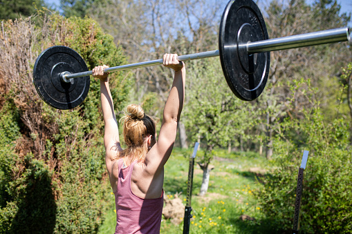 A fit young woman is doing a barbell press in her back yard on a sunny day