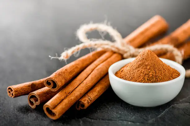 Cinnamons dried sticks on dark stone table. Cinnamon ginger powder in white bowl on black board.