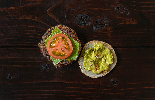 An overhead, table top display of a wild rice, mushroom, black bean vegan burger with vegan mayo, romaine lettuce, tomato and guacamole on a bun, set on top of a rustic wooden surface.