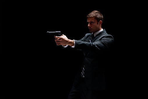 One person of aged 20-29 years old with short hair caucasian young male business person in front of black background wearing businesswear who is agressive who is violence and holding weapon and using pistol