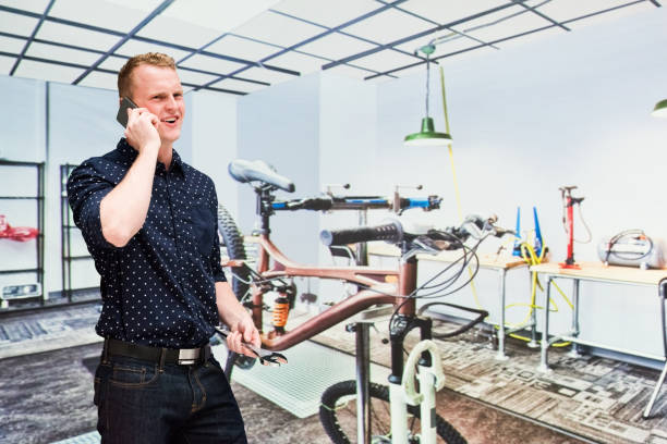 young male sales occupation at the bicycle shop in front of merchandise in the sports shop wearing jeans and holding wrench and using smart phone - sales clerk store manual worker retail imagens e fotografias de stock