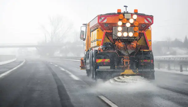 Photo of Snow plow salting street in winter time. Orange truck deicing.