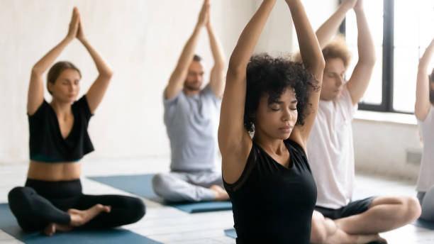 African trainer and group of people meditating during yoga class During morning routine yoga session after work out diverse people relaxing seated cross-legged practising meditation closed eyes raised arms up performing Namaste gesture, focus on mixed-race trainer spiritual enlightenment stock pictures, royalty-free photos & images