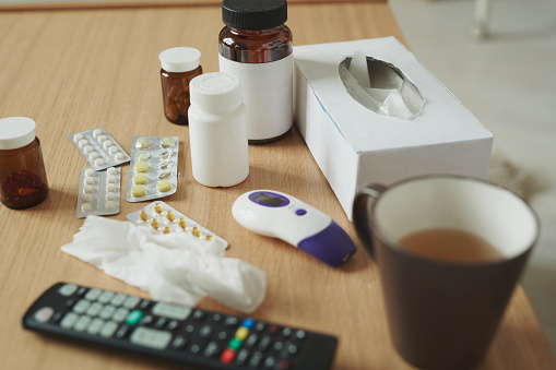 Number of plastic and glass bottles with pills and blisters with tablets, paper tissues, thermometer, mug with hot drink and remote control on table