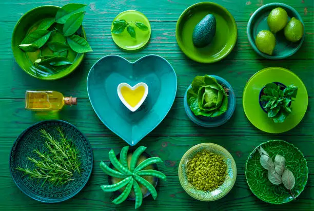 Home inventory all in green crockery plates with vegetarian food and heart shape olive oil on green wooden background monochromatic green