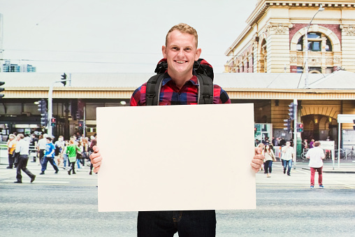 Waist up of with redhead caucasian male who is outdoors wearing plaid shirt who is showing cool attitude who is showing with hand and holding sign with copy space