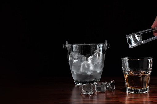 hand with tongs holding ice-cube above a glass with whiskey, ice bucket behind the alcoholic drink on wooden table in restaurant. Black background for copy space