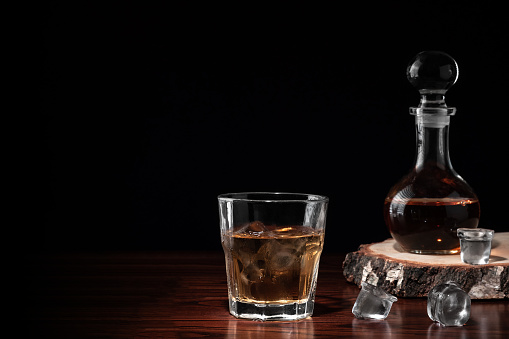 Crystal decanter or carafe of whiskey on cross section of birch tree trunk and glass with alcoholic drink and ice on wooden table in restaurant. Black background for copy space