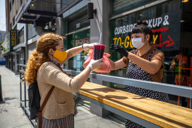 Woman Wearing Homemade Mask Picks Up Food at Restaurant During Covid-19 Lockdown A Mexican restaurant adapts to the Covid-19 lockdown. The owner hands an order to a customer outside the restaurant; they are both wearing gloves and masks. lockdown business stock pictures, royalty-free photos & images