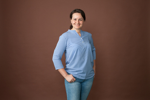studio close-up portrait of an attractive 30 year old woman with brown ponytail hair in a blue blouse and blue jeans on a brown background