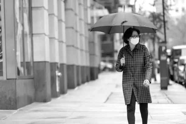 Senior Asian woman walking in the rain on city street. Senior adult wearing surgical mask and practicing social distancing on the sidewalks of Manhattan, New York during coronavirus pandemic.