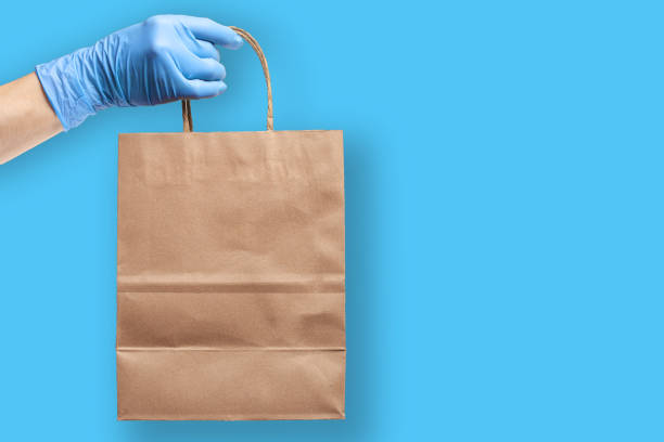 Hand wearing rubber protective glove   holds paper grocery bag. Grocery shopping and delivery concept during novel virus pandemic. Protection equipment used while shopping. stock photo