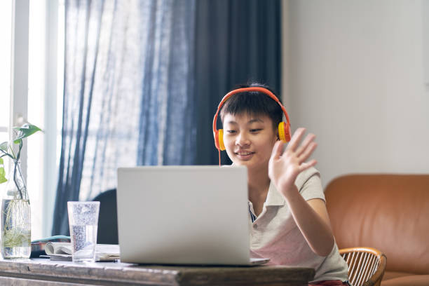 Asian teenage boy studying at home wearing headset, smiling & say hello stock photo