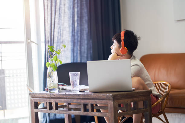 Asian teenage boy looking out of window when studying at home wearing headset stock photo