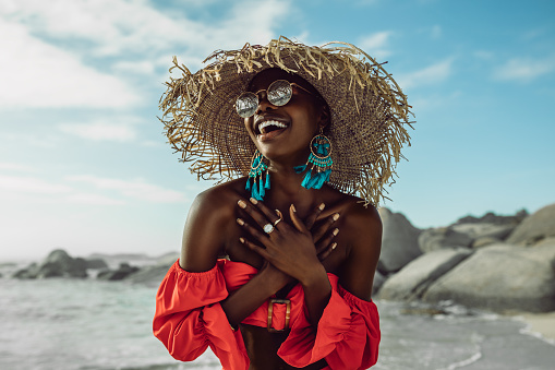 Beautiful african woman in red dress wearing straw hat smiling on the beach. Female in sundress enjoying on the beach.