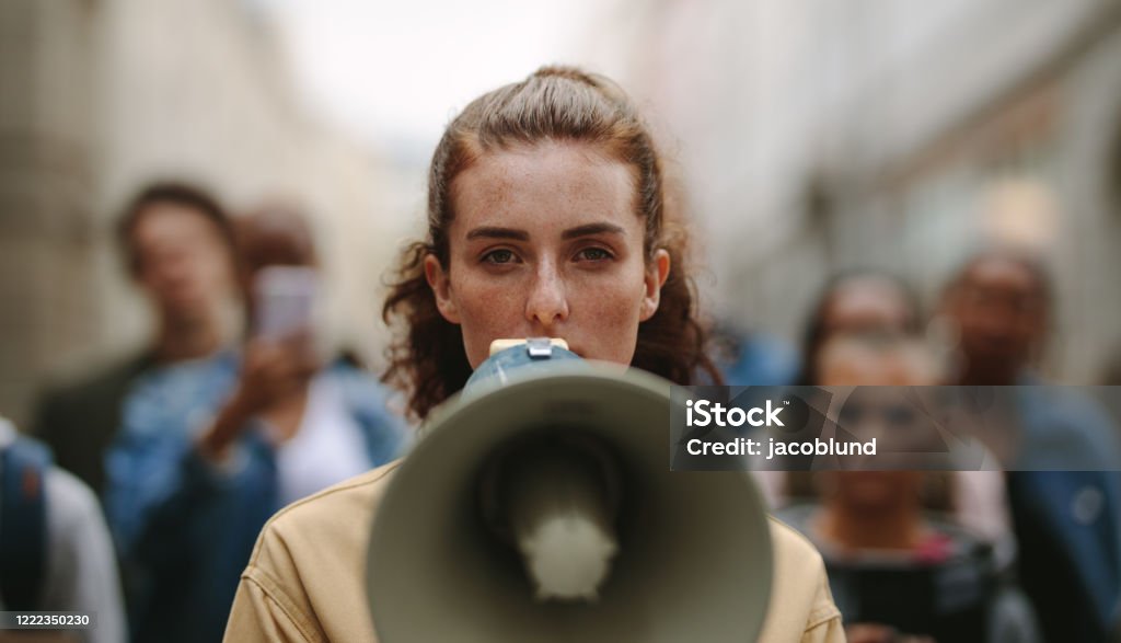 Female activist protesting with megaphone during a strike Female activist protesting with megaphone during a strike with group of demonstrator in background. Woman protesting in the city. Megaphone Stock Photo