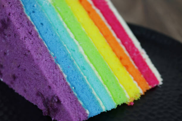 Image of slice of rainbow cake showing multi-coloured sponge layers photo with butter icing cream on black dimpled tray