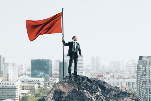Winner businessman with red flag standing on mountain. City skyline background. Leadership and success concept