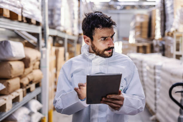 istock Young worker in sterile protective suit standing in warehouse with hand on hip and holding tablet while looking at camera. He checking out which sector is disinfected from corona virus. 1222372916